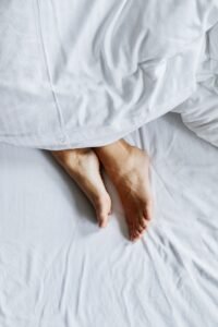 Pés descalços sob um edredom branco na cama, contexto de conforto e cuidado com a pele dos pés