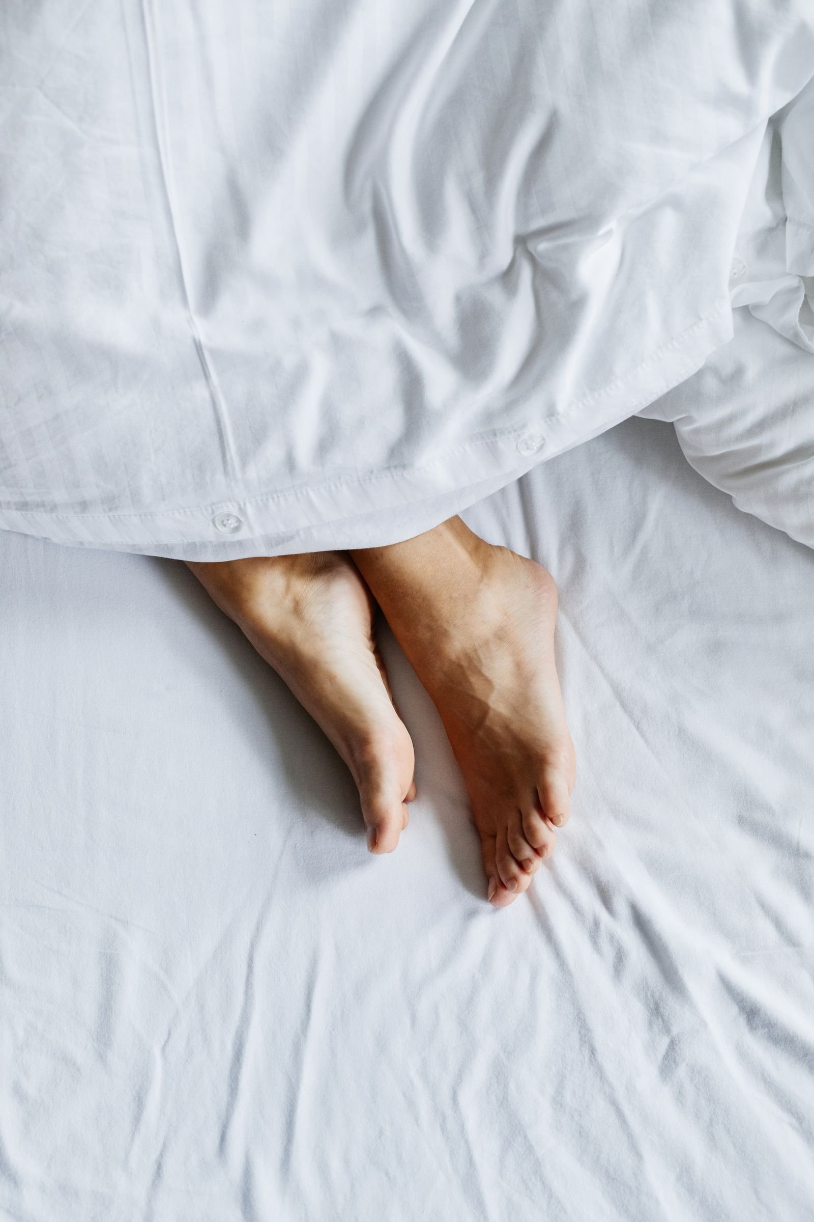 Pés descalços sob um edredom branco na cama, contexto de conforto e cuidado com a pele dos pés